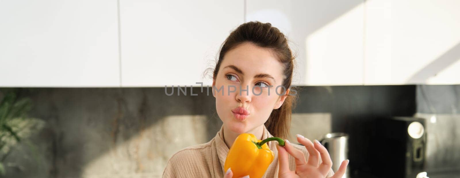 Close up of brunette woman in robe, holding yellow pepper, thinking what to cook, preparing salad or soup, making vegetarian meal with vegetables, standing in kitchen by Benzoix