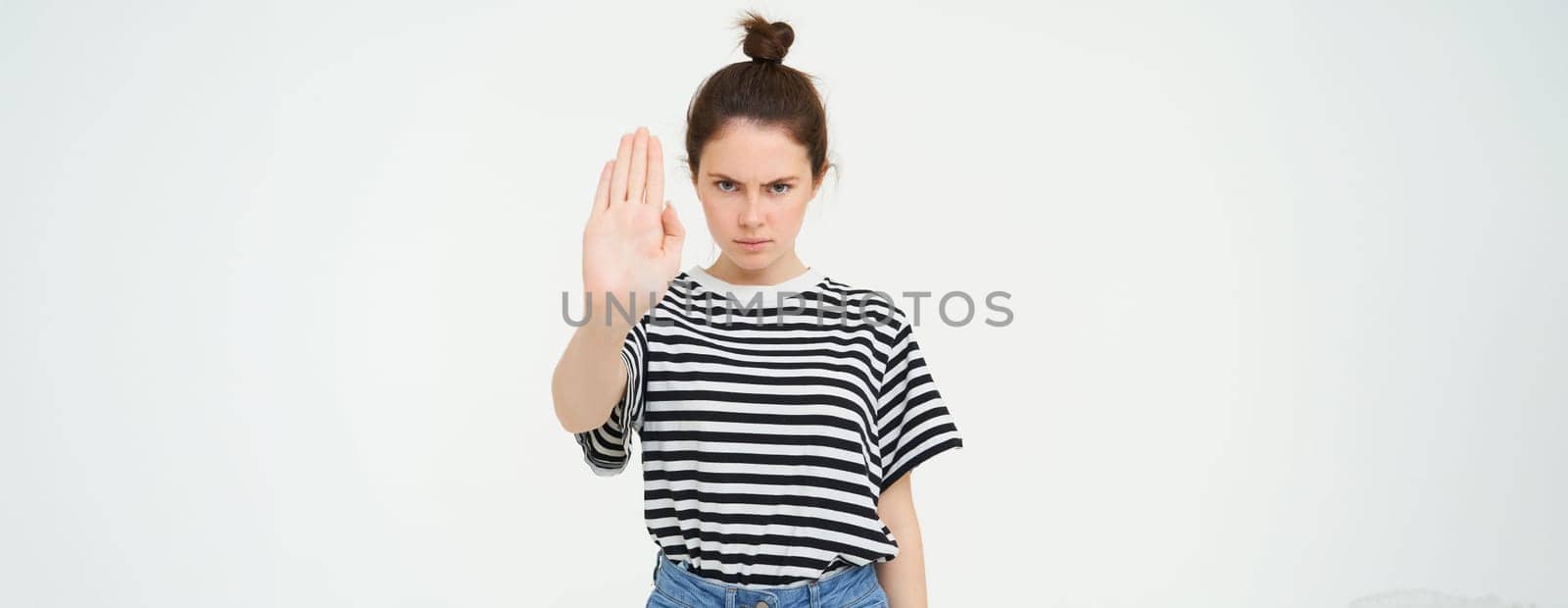 Image of serious and confident young woman, shows stop sign, taboo gesture, prohibit something bad, standing over white background.