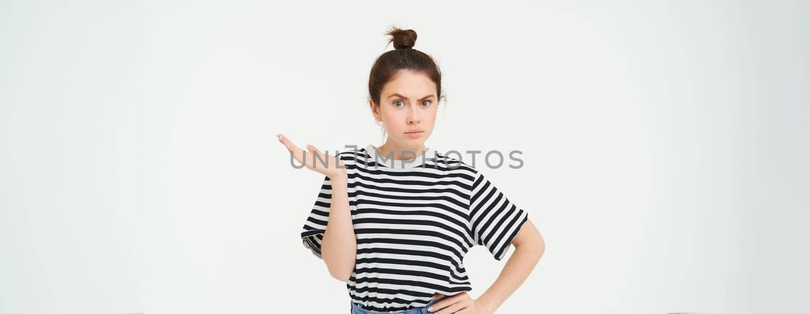 Portrait of confused girl, shrugging should and raising one hand up, looks puzzled, cant understand something, standing over white background.