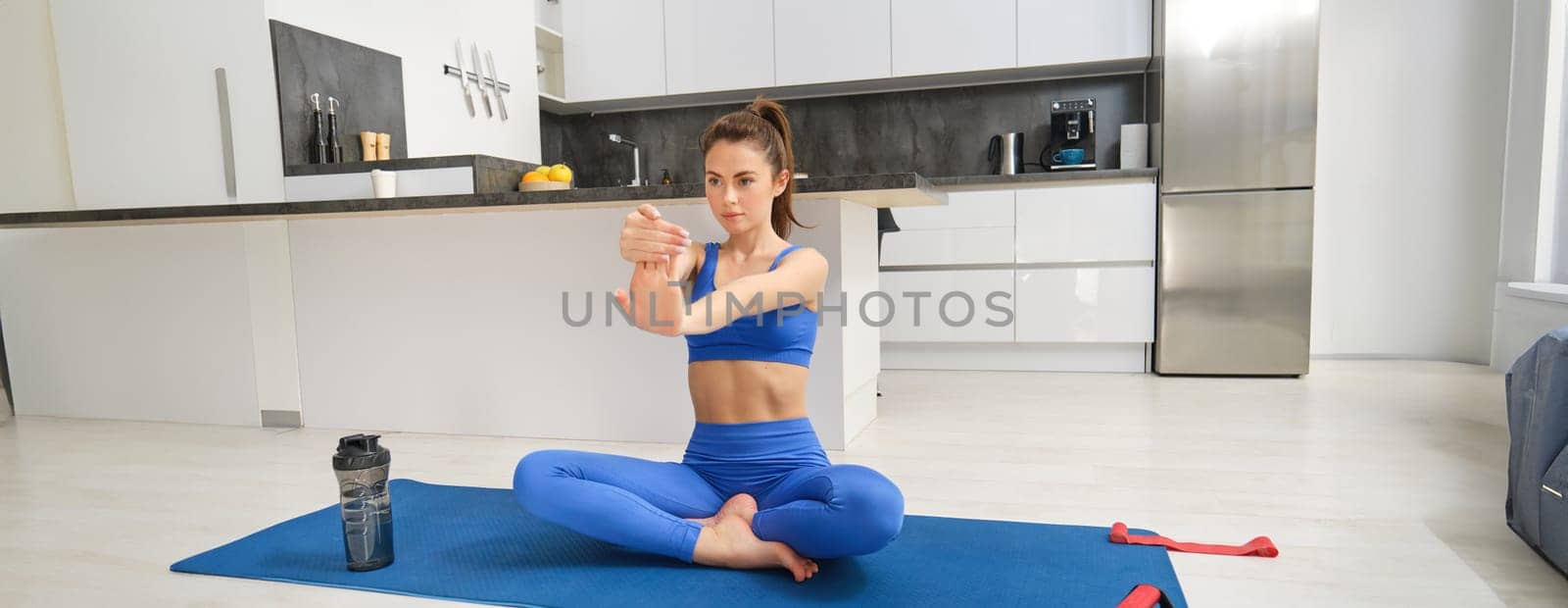 Young woman does workout from home, sits on yoga mat and stretches her hands, fitness training session in living room.
