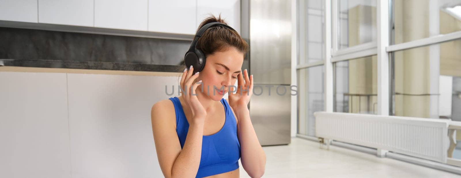 Young woman puts on wireless headphones to listen music during workout at home, wears sportsbra, smiles with pleased face by Benzoix