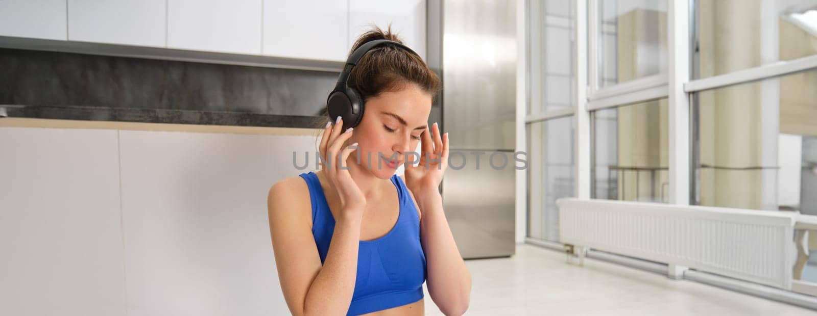 Young woman puts on wireless headphones to listen music during workout at home, wears sportsbra, smiles with pleased face.