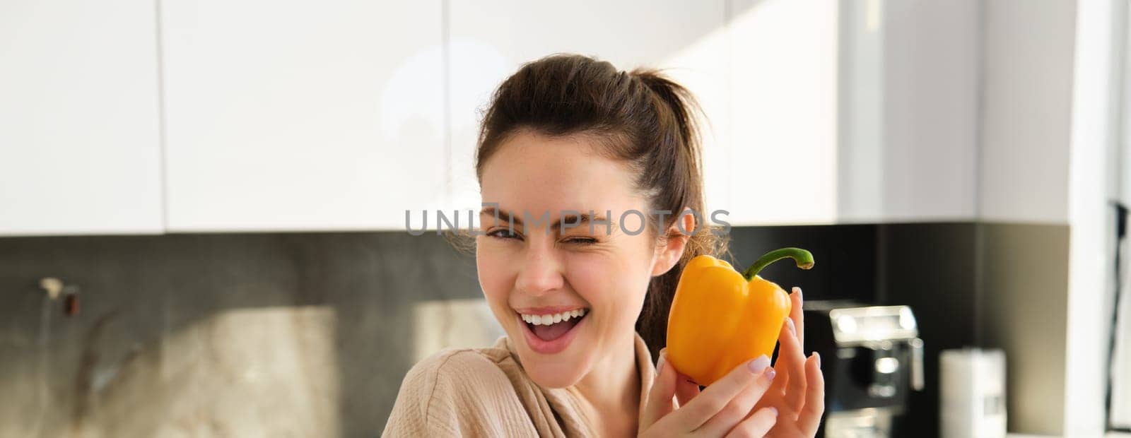 Close up portrait of cute winking girl, showing yellow pepper, standing in kitchen, making vegetarian meal, making salad, eating healthy vegetables by Benzoix