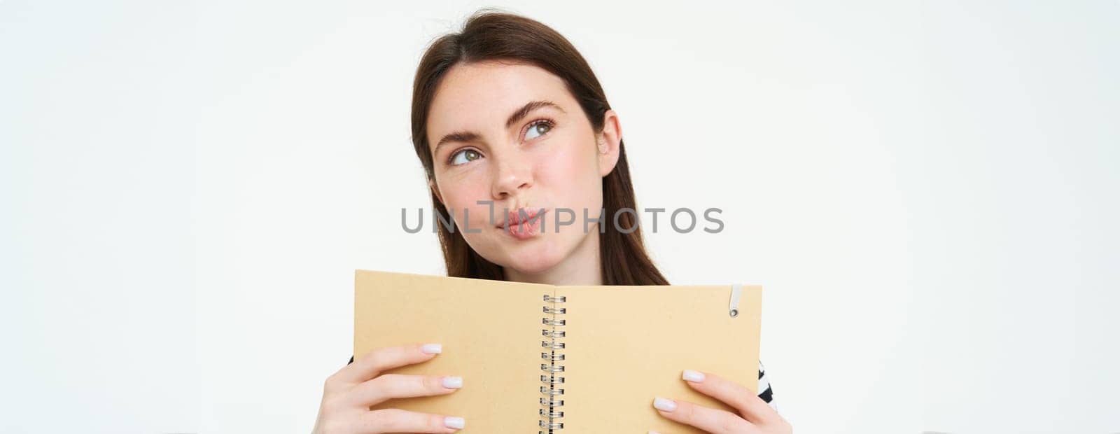 Portrait of young woman makes notes in her planner, writes in diary, checks her schedule in notebook, holding organizer in hands, stands over white background by Benzoix