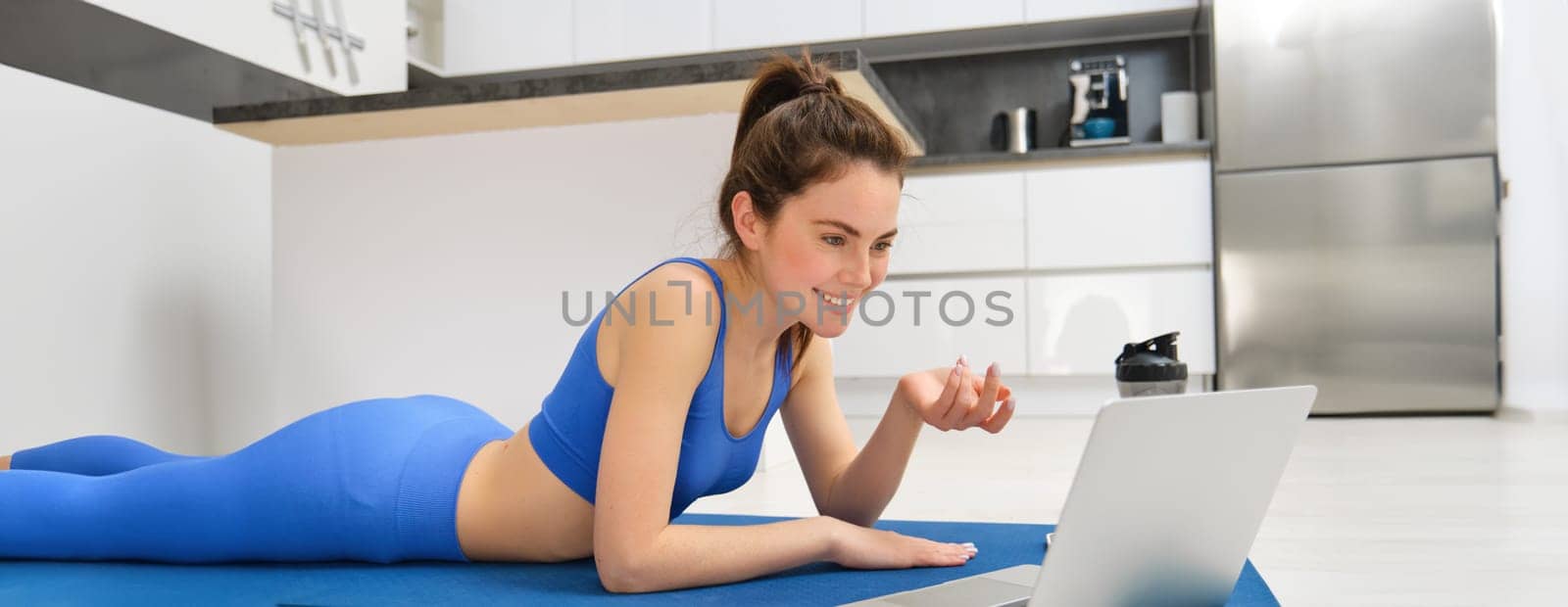 Image of young fitness girl talking online on laptop, video chats, laying on floor rubber mat, doing exercises, discuss workout training, yoga from home.