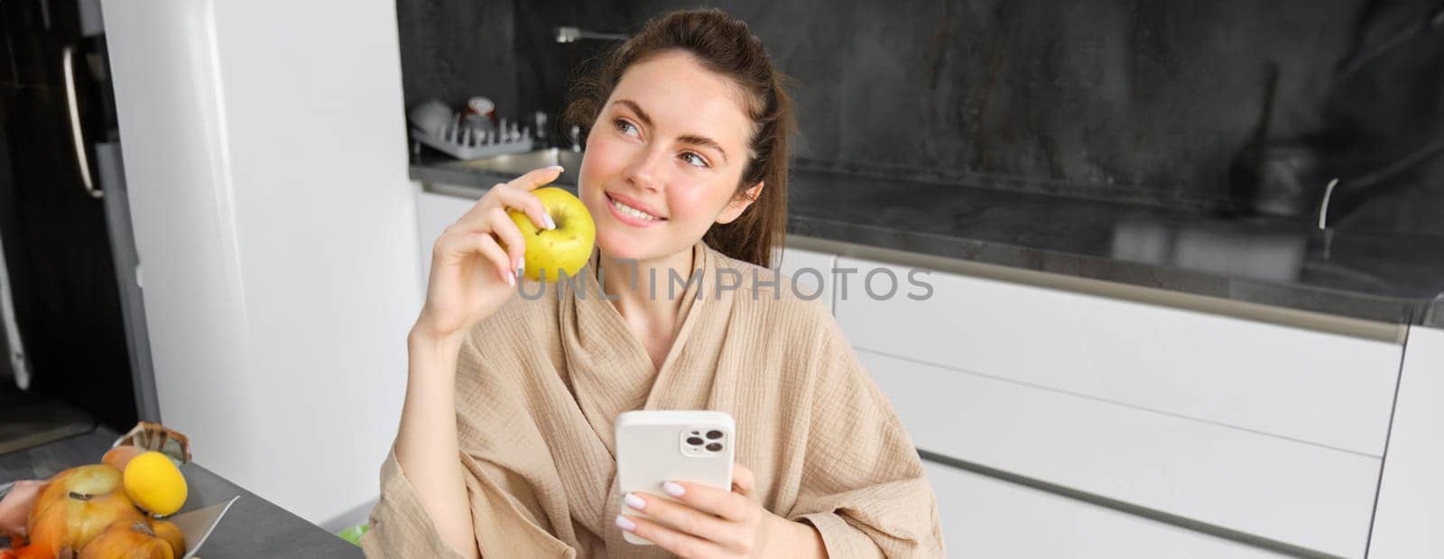Close up portrait of happy young woman in bathrobe, sitting in the kitchen and using mobile phone, holding an apple, order fruits and vegetables online, using smartphone app for groceries delivery.