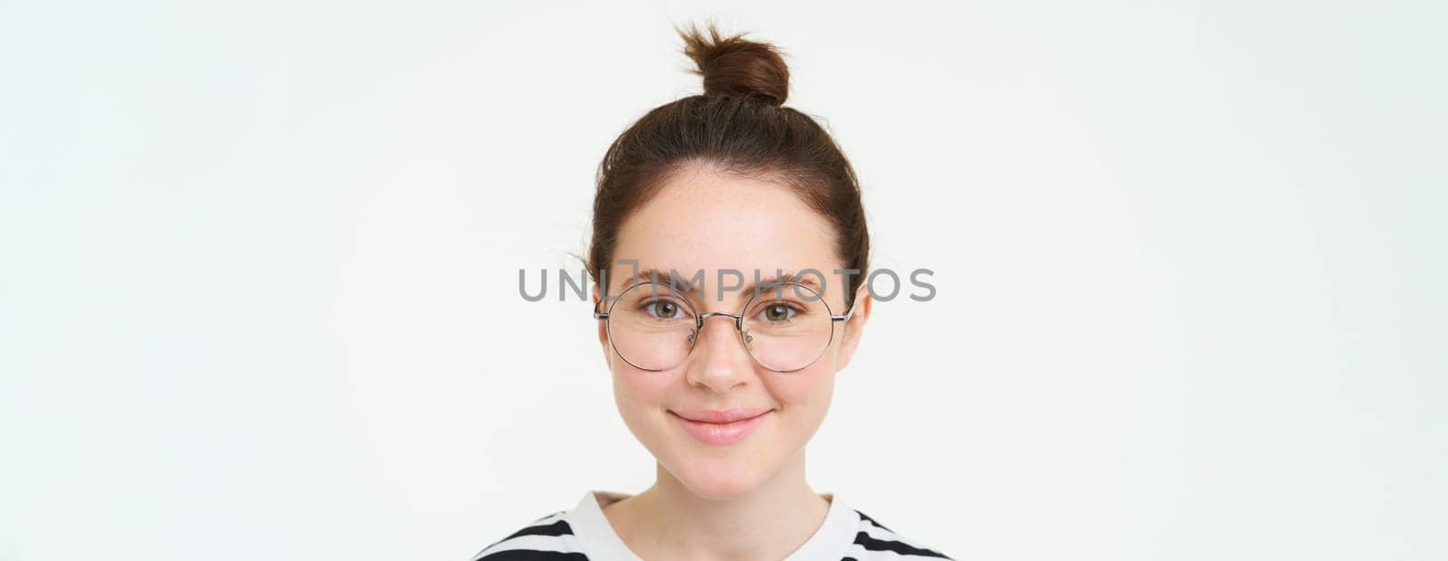 Close up portrait of cute young woman in glasses, looking at camera and smiling, trying on new spectacles, standing over white background by Benzoix