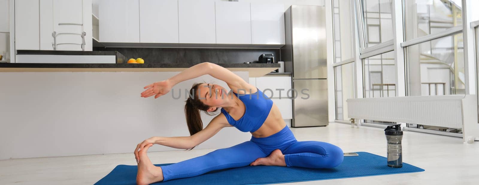 Portrait of beautiful young woman gymanst, stretching her body during workout at home, doing fitness exercises, stretching legs and arms, wearing blue sportsbra and leggings.