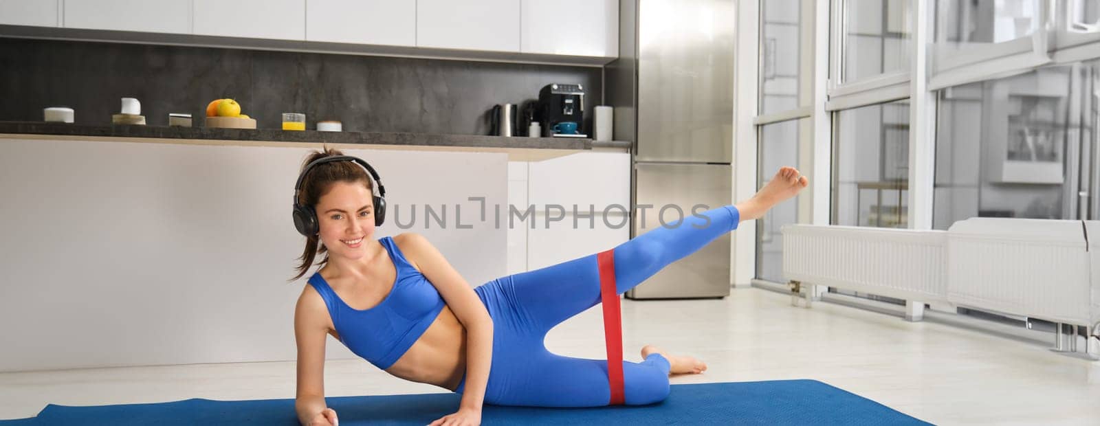 Workout and fitness concept. Woman in headphones does exercises at home, stretching rubber resistance band with legs, laying on yoga mat in living room, lifting her foot up.
