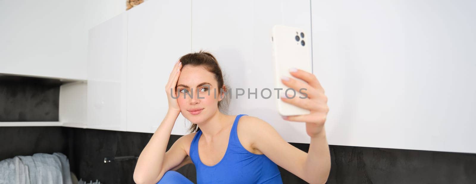 Portrait of beautiful sportswoman, wearing sportsbra and leggings, sitting in kitchen and taking selfie, posing for photo on smartphone app.