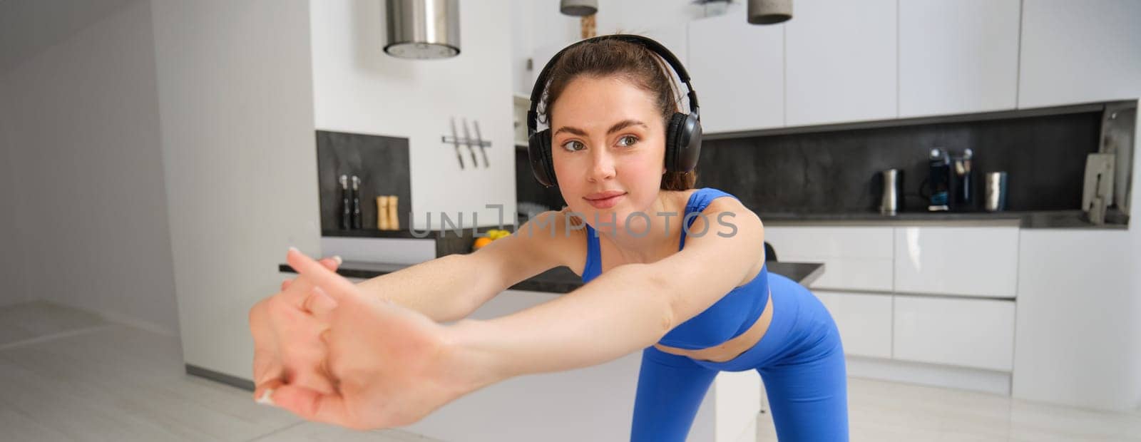 Close up portrait of fit and healthy woman, smiling while doing workout exercises, listening music in wireless headphones, stretching arms, aerobics training at home, wearing blue activewear.