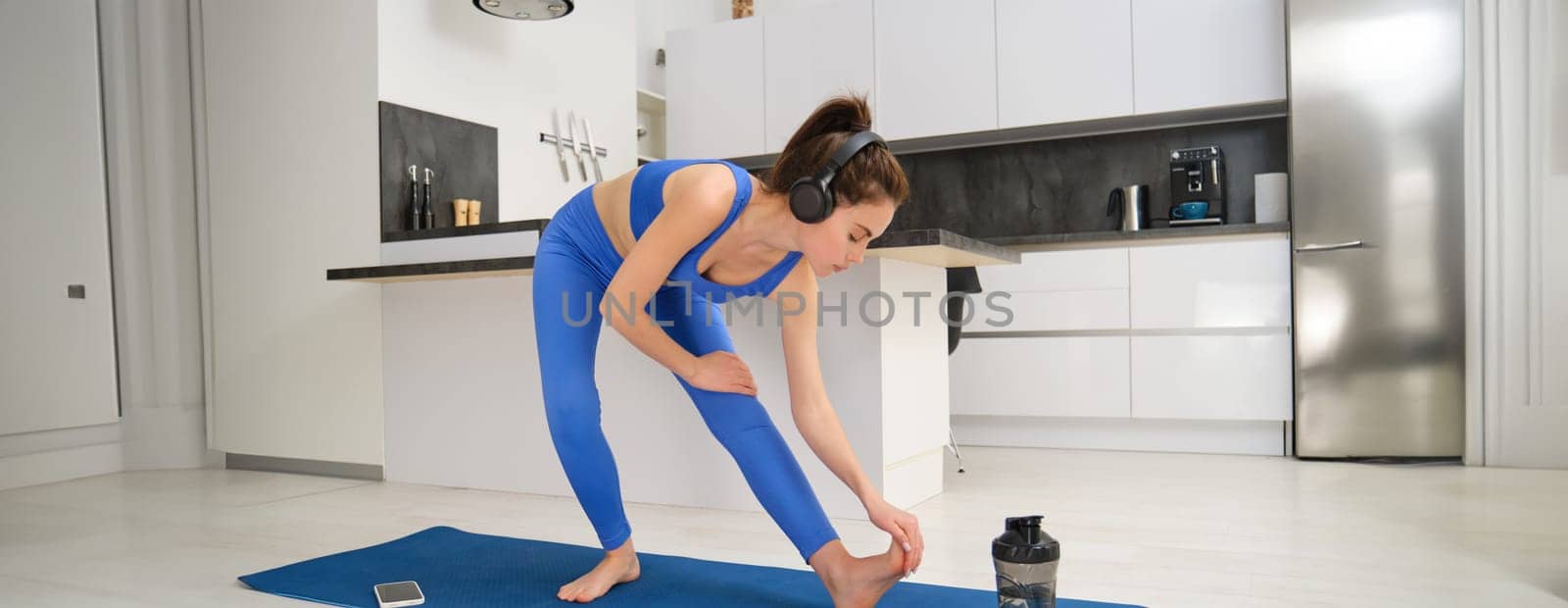 Image of young fit woman doing fitness workout from home, stretching her legs, warm-up before aerobics training, listening music in wireless headphones, exercising in living room.