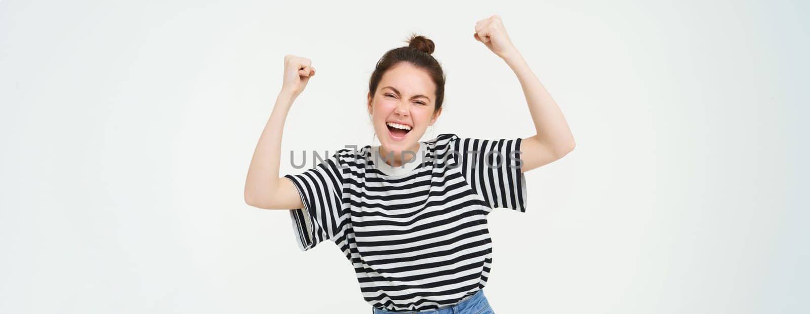 Portrait of happy young sports girl, fan rooting for team, celebrating victory, raising hands up, chanting, triumphing, standing over white background. Copy space