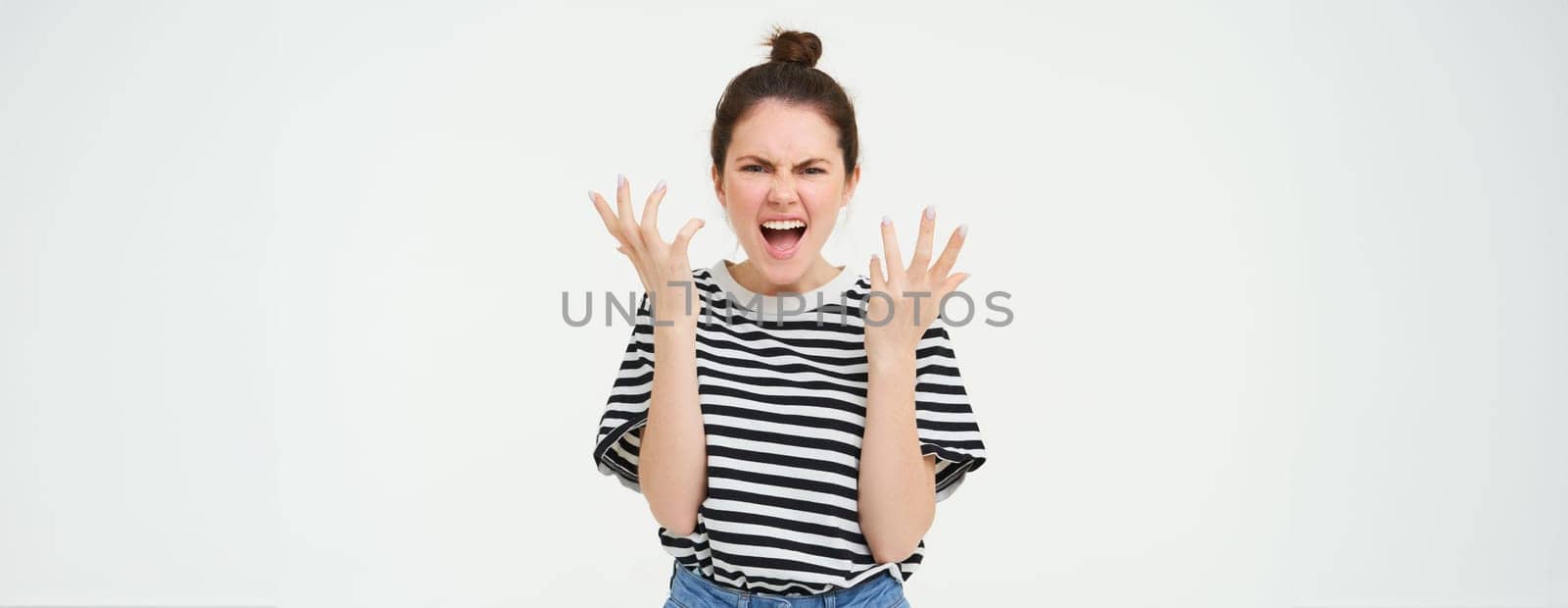 Image of angry woman shouts and shakes hands, stands in casual clothes over white background. Copy space