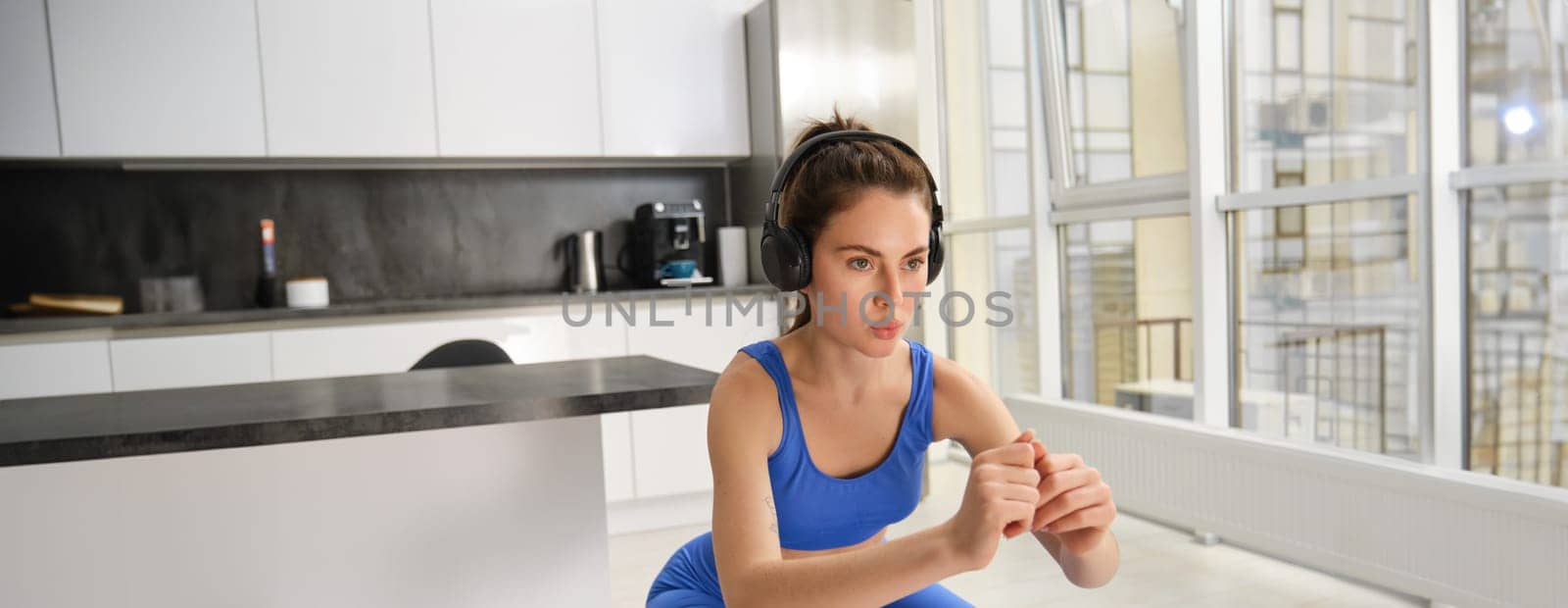 Image of young sportswoman, fitness instuctor doing squats workout at home, listening music in wireless headphones and stretching arms, doing exercises in living room.