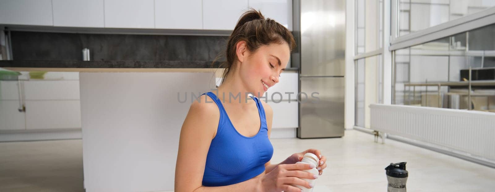 Image of young woman, sporty fitness girl taking vitamins, dietary supplements for muscles strength, sitting on yoga mat and workout.