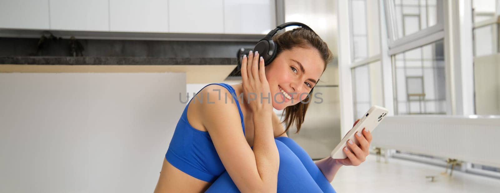 Fitness and wellbeing. Portrait of young woman doing fitness exercises, workout from home, wearing headphones and using smartphone, exercising indoors.