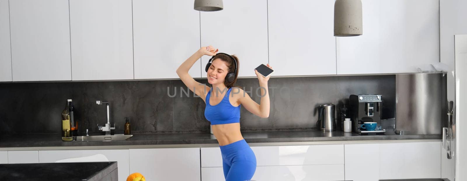 Portrait of beautiful fitness blogger, woman in headphones, listening music and dancing in kitchen, wearing blue leggings and sportsbra.