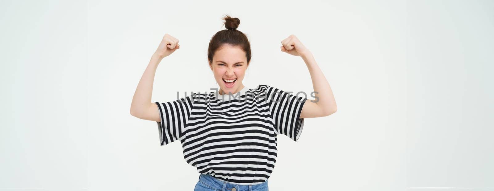 Image of sassy, strong and confident woman shows her muscles, flexing biceps, raising her arms high, standing over white background.