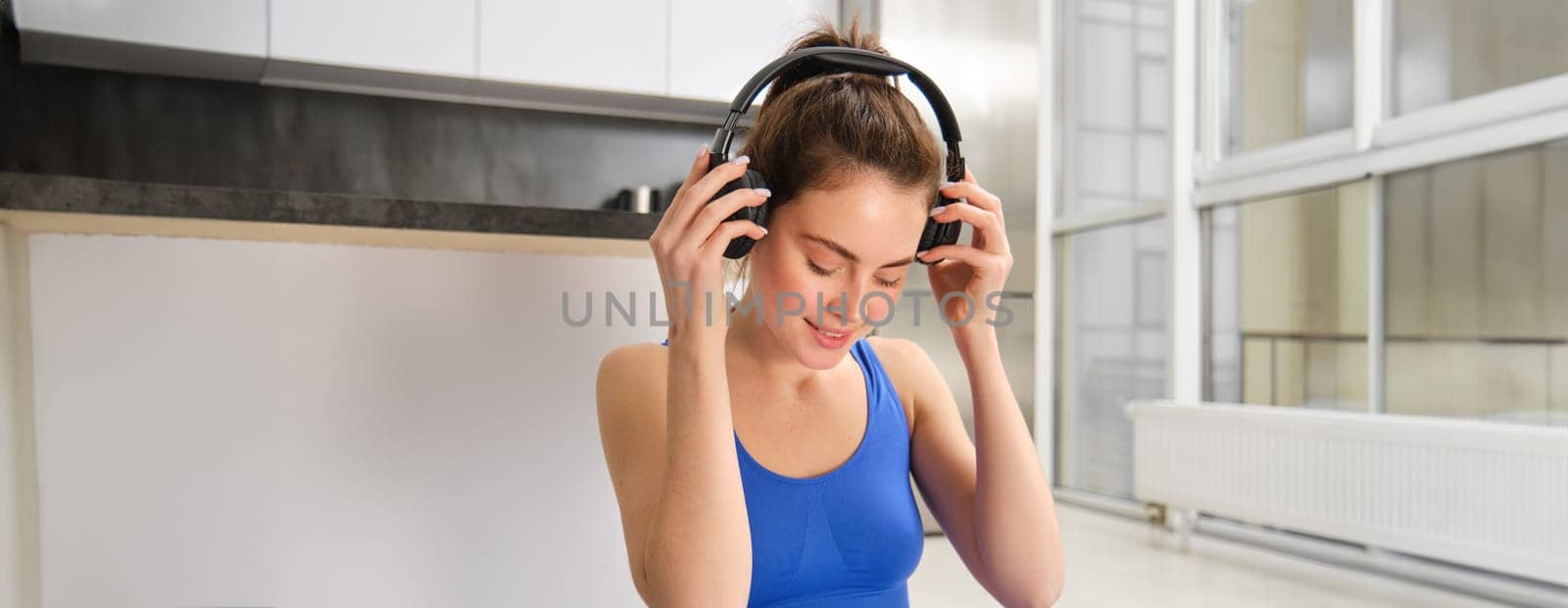Smiling fitness woman puts on wireless headphones before her home workout, yoga training in blue tracksuit.