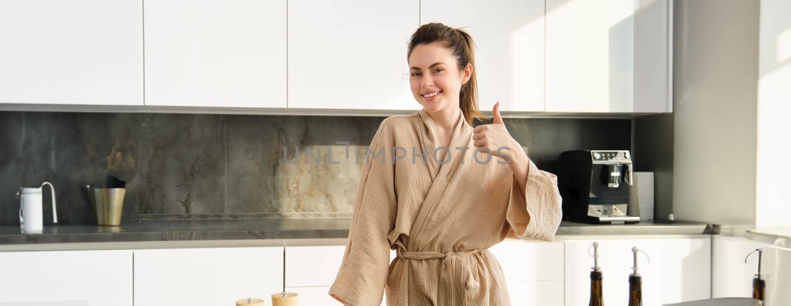 Portrait of smiling woman in bathrobe, shows thumbs up, stands in kitchen, cooking salad, preparing vegetarian dinner, chopping vegetables on board by Benzoix