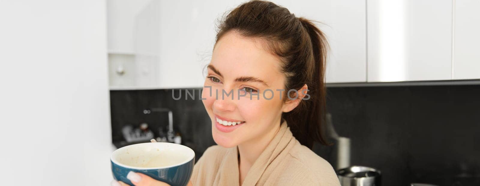 Portrait of good-looking young woman starting her day with cup of coffee, standing in the kitchen and drinking cappuccino from big mug, enjoying favourite drink in the morning by Benzoix