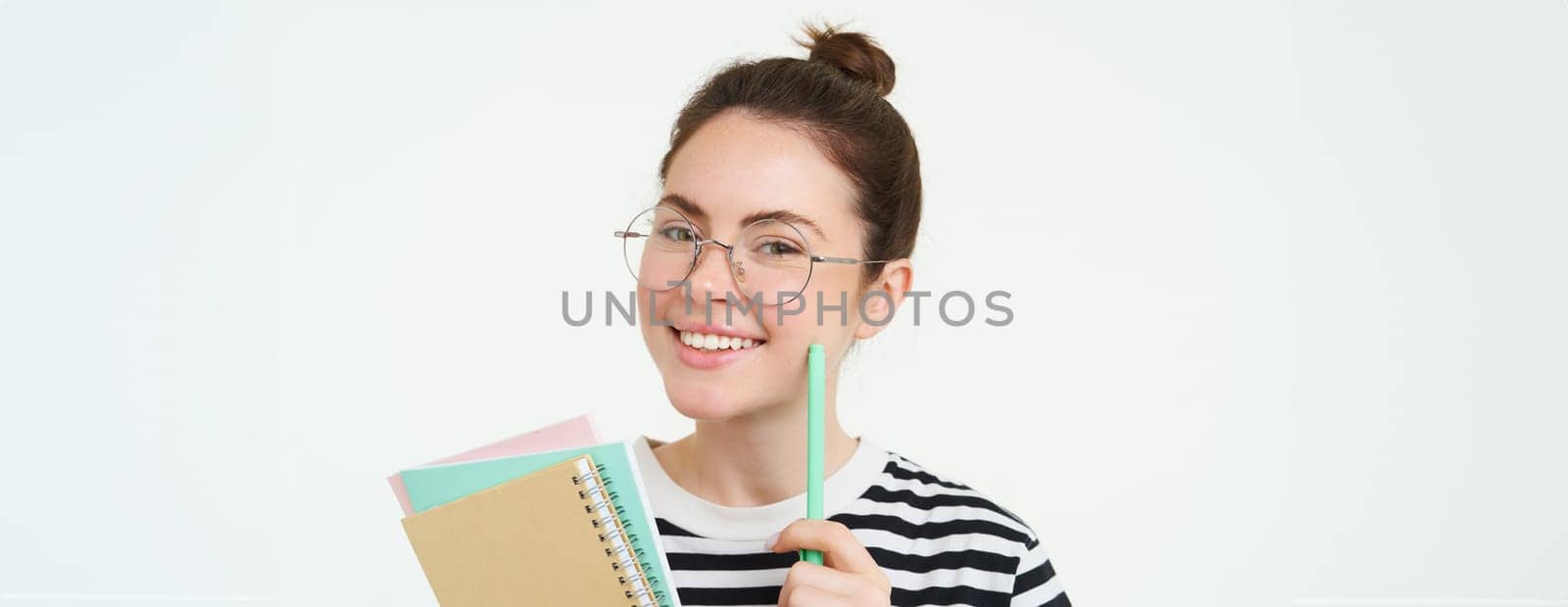 Portrait of smart girl in glasses, tutor holding pen and notebooks, student carry her homework notes, standing over white background by Benzoix