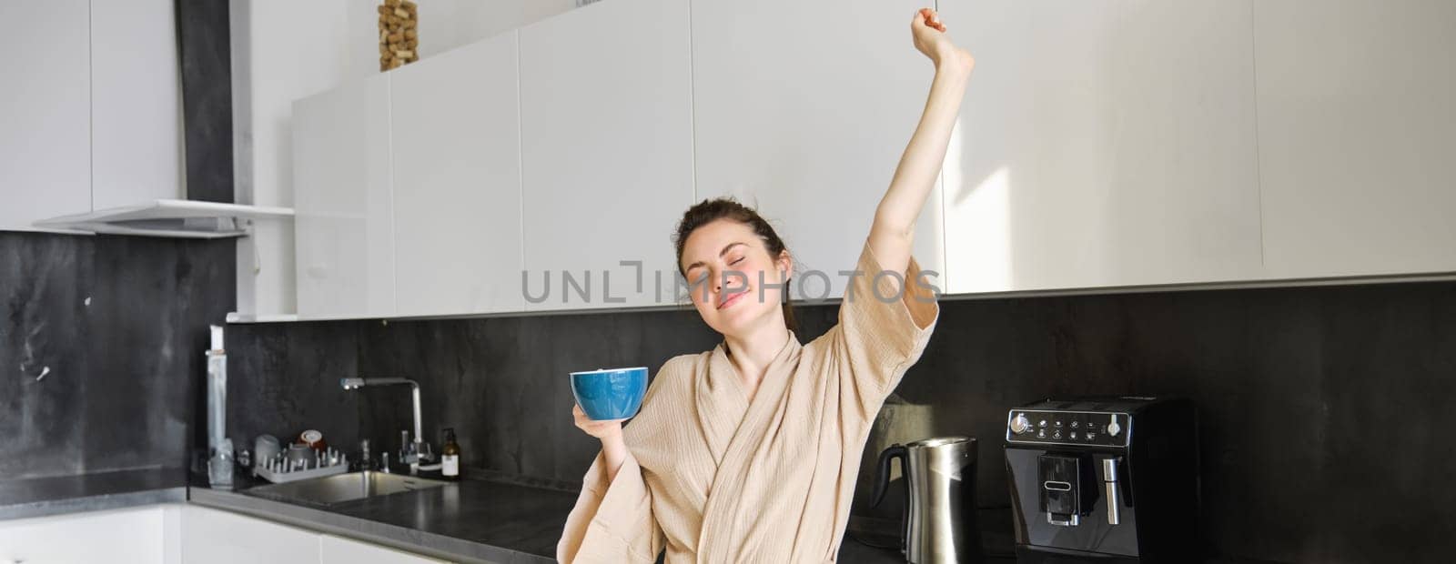 Portrait of carefree brunette woman, dancing and stretching hands in the kitchen, drinking coffee, smiling pleased in the morning by Benzoix