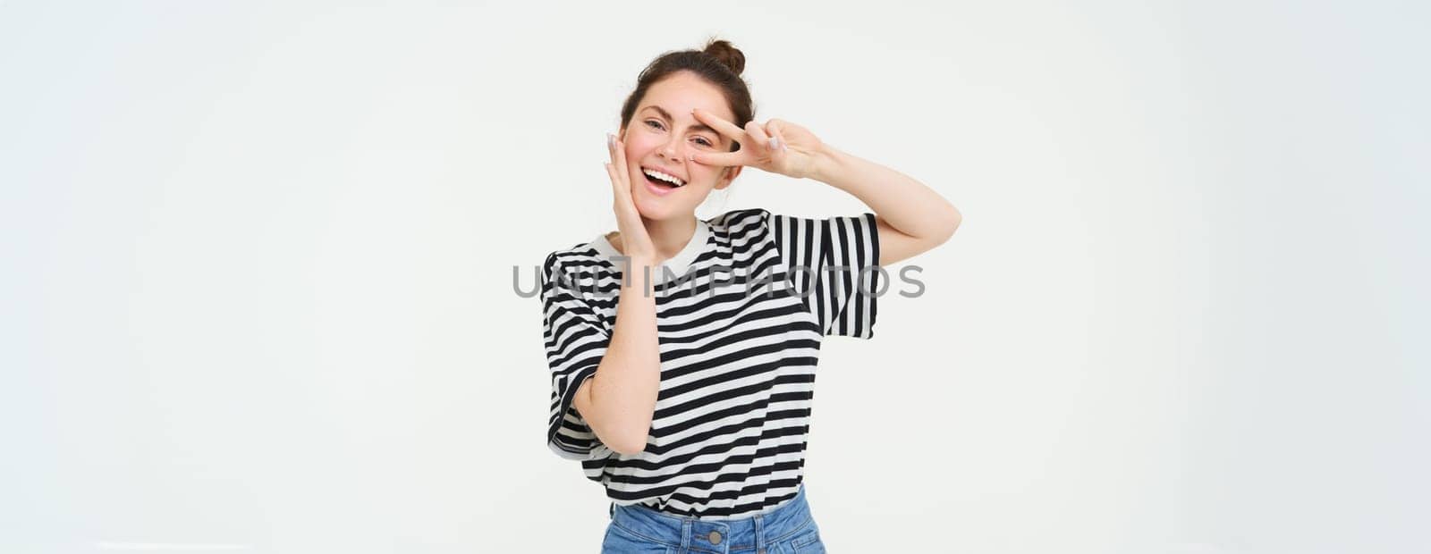 Cute european woman posing for photo, holding peace, kawaii hand gesture near face, smiling at camera, standing over white background.