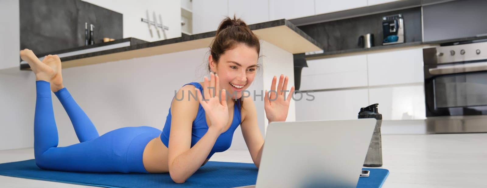 Portrait of beautiful, fit young woman on floor at home, saying hello at laptop screen, waving hand and smiling, workout remotely with friend, doing yoga class online by Benzoix