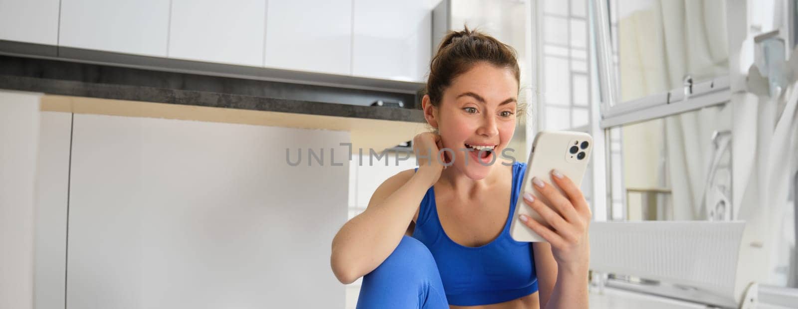 Close up shot of young happy woman, doing sports, workout at home, checking her phone, looking amazed at smartphone screen by Benzoix