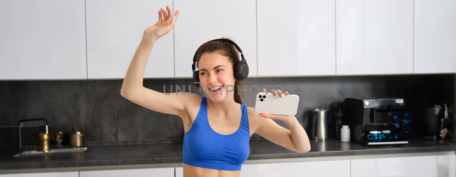 Portrait of beautiful sports woman, wearing activewear, dancing in kitchen with headphones, listening music and having fun, feeling energy after workout session.