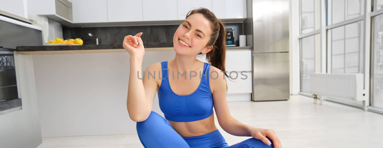 Young sporty girl in living room, sits on yoga mat, smiles and looks at camera, fitness instructor does online training from her house, stretches, workout at home.