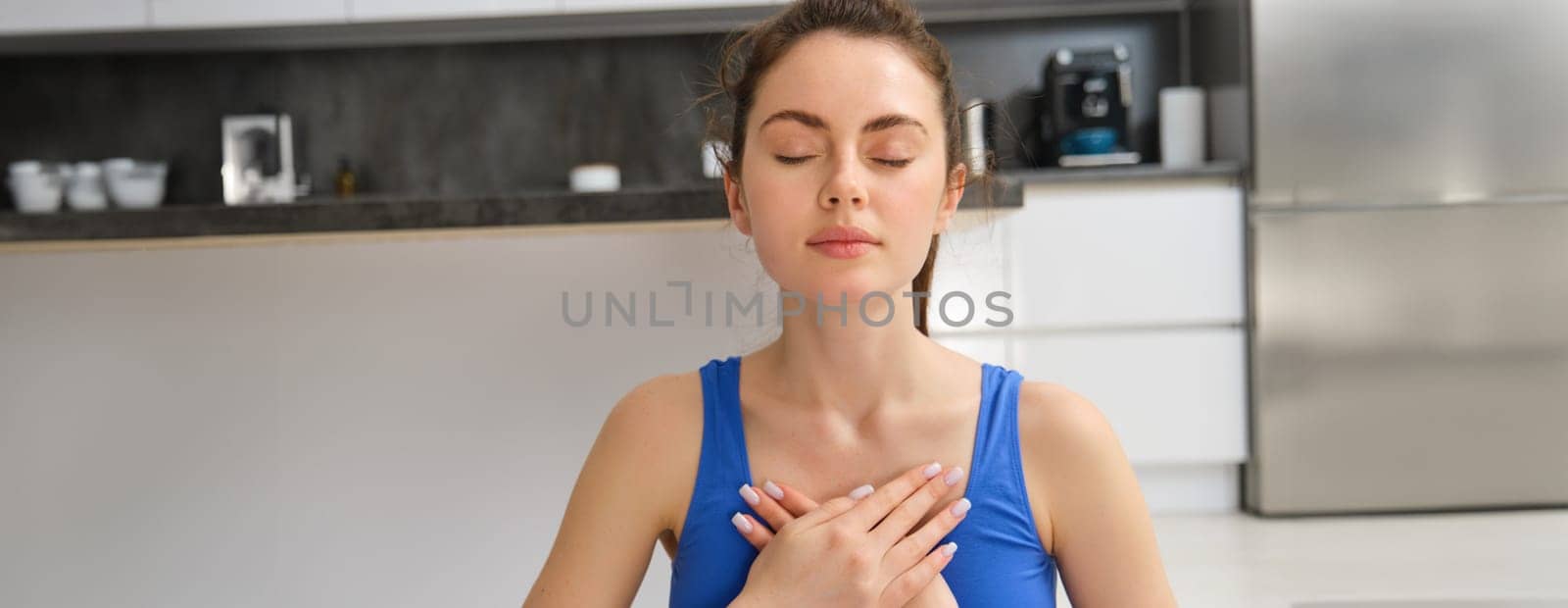 Image of calm and relaxed woman meditating, doing breathing practices, holding hands on chest during yoga session at home. Sport and mindfulness concept