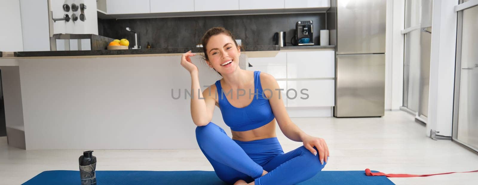 Image of young sport woman sitting at home on yoga mat, doing workout, stretching fitness exercises on floor in living room, smiling and looking happy at camera.