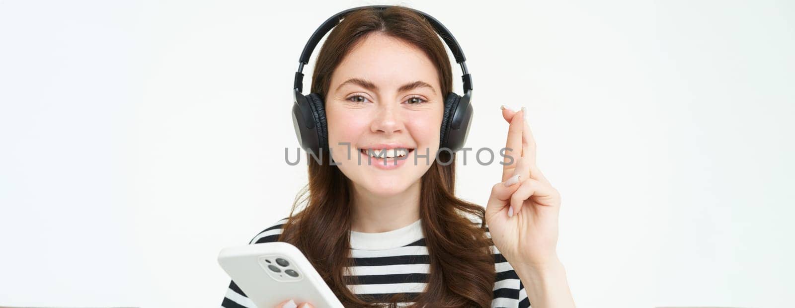 Portrait of smiling female model using headphones and smartphone, hold fingers crossed, makes wish, hopes for something, anticipates, stands over white background.