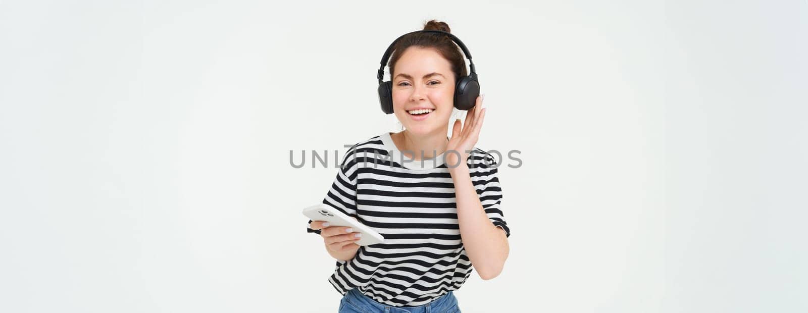 Portrait of happy woman with smartphone changes song on mobile phone streaming app, listens music in headphones, dancing against white background by Benzoix