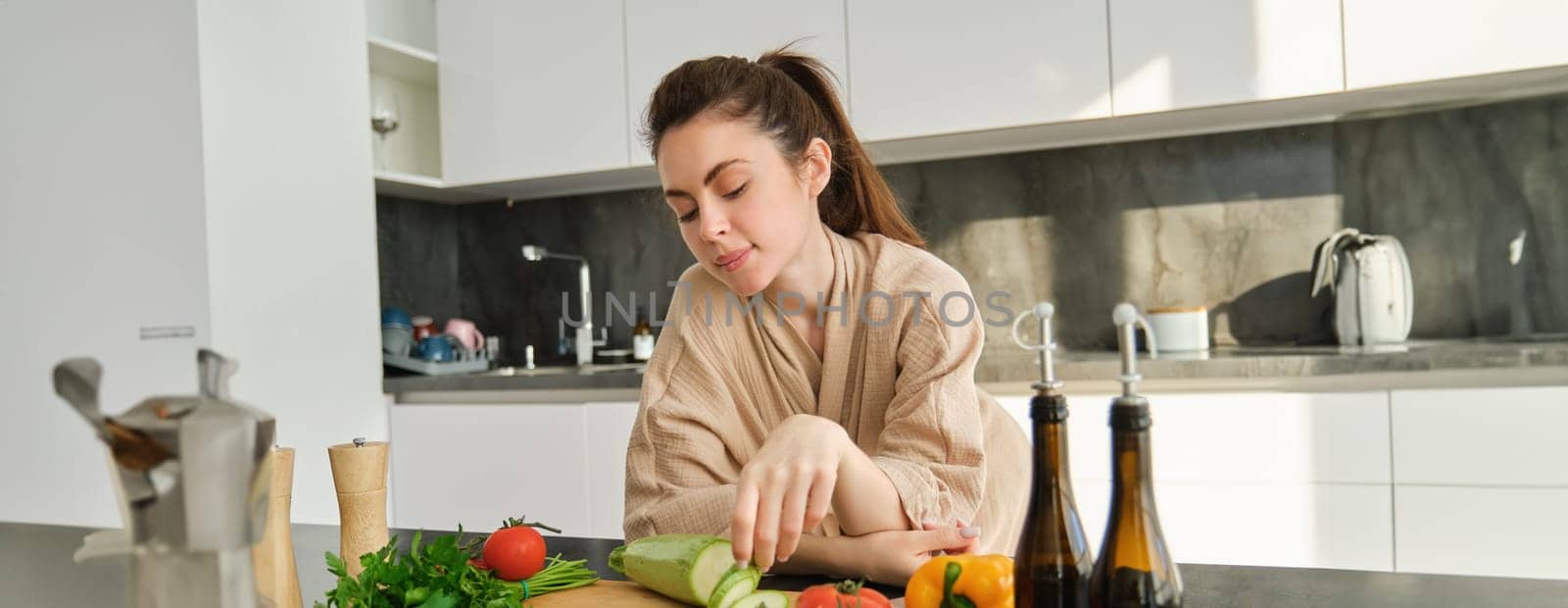 Portrait of cute young woman in bathrobe, cooking meal, standing near chopping board with vegetables, holding zucchini, making salad or vegetarian dinner by Benzoix