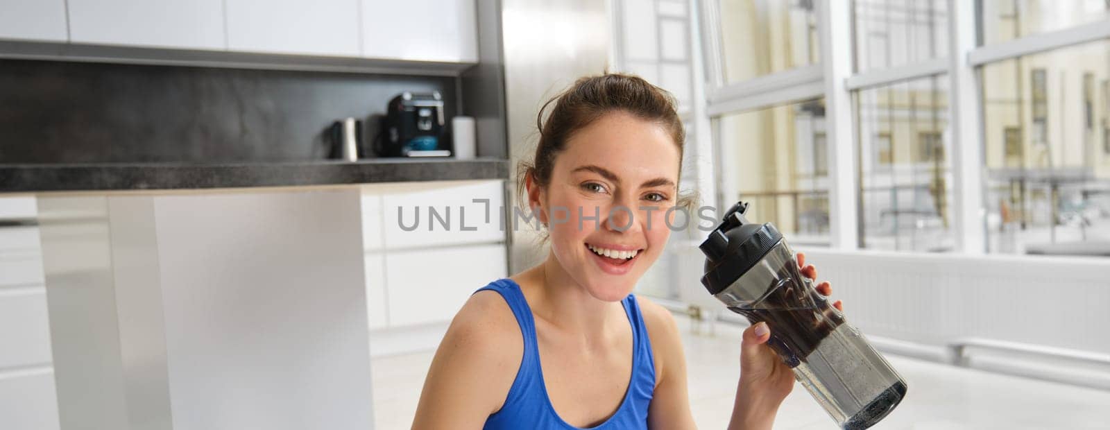 Beautiful young sportswoman drinks water, stays hydrated during workout training at home, doing fitness exercises in living room.