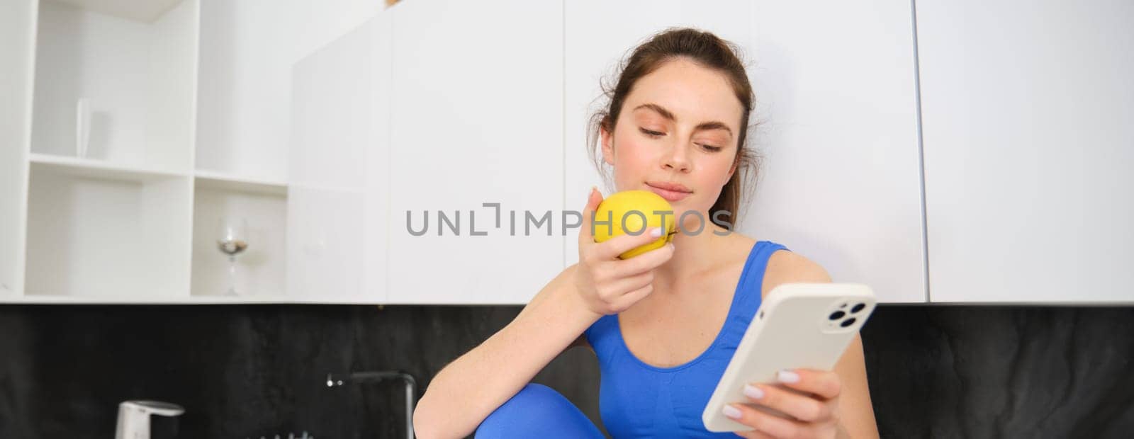 Image of stylish sportswoman, young fitness instructor sitting in kitchen and eating an apple, holding smartphone, using social media app on mobile phone by Benzoix