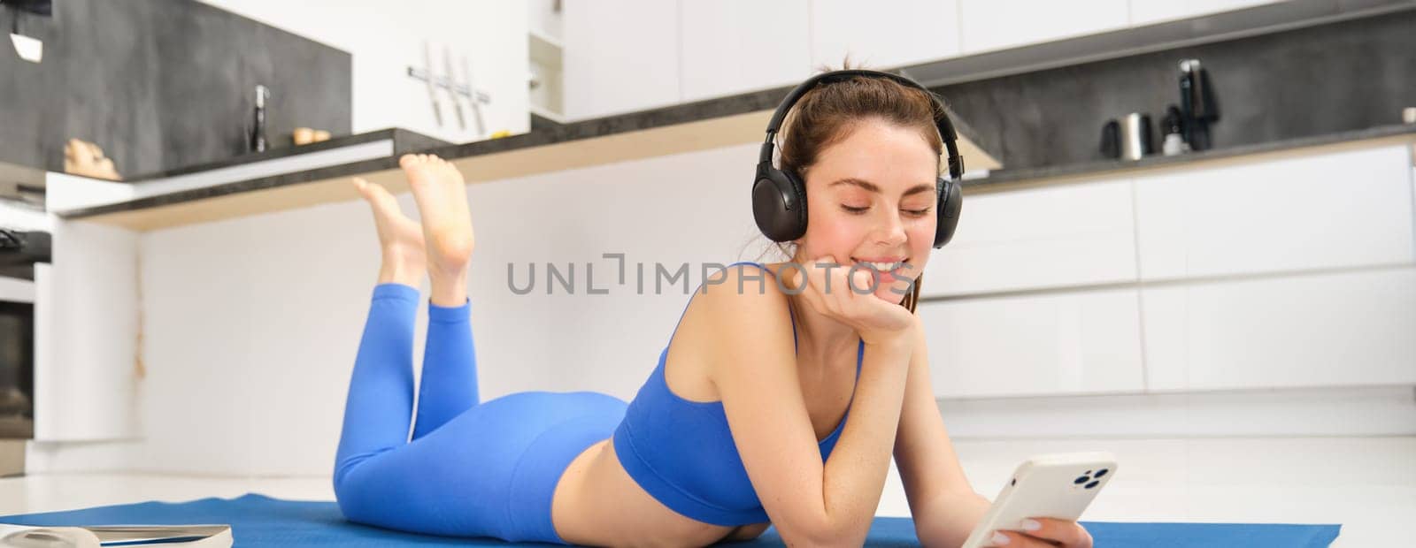 Portrait of fitness woman, listening yoga music in headphones, using her smartphone during workout at home, laying on blue sports mat.