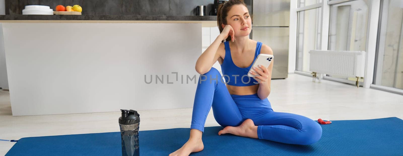 Indoor shot of young sportswoman holding smartphone, sits with fitness water bottle, uses sports app to workout, do yoga from home by Benzoix