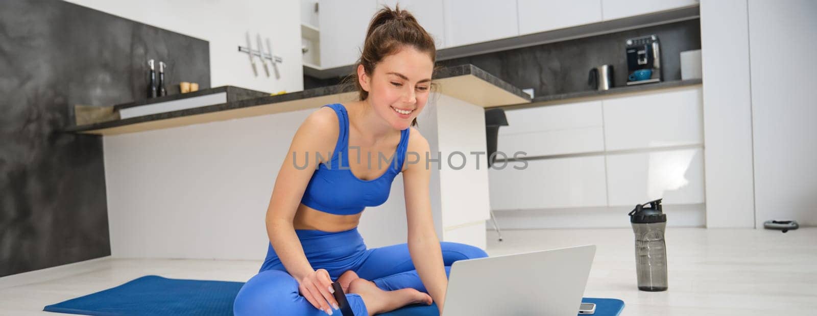Portrait of young woman following online fitness lesson at home, sitting on yoga mat and using laptop during aerobics workout by Benzoix