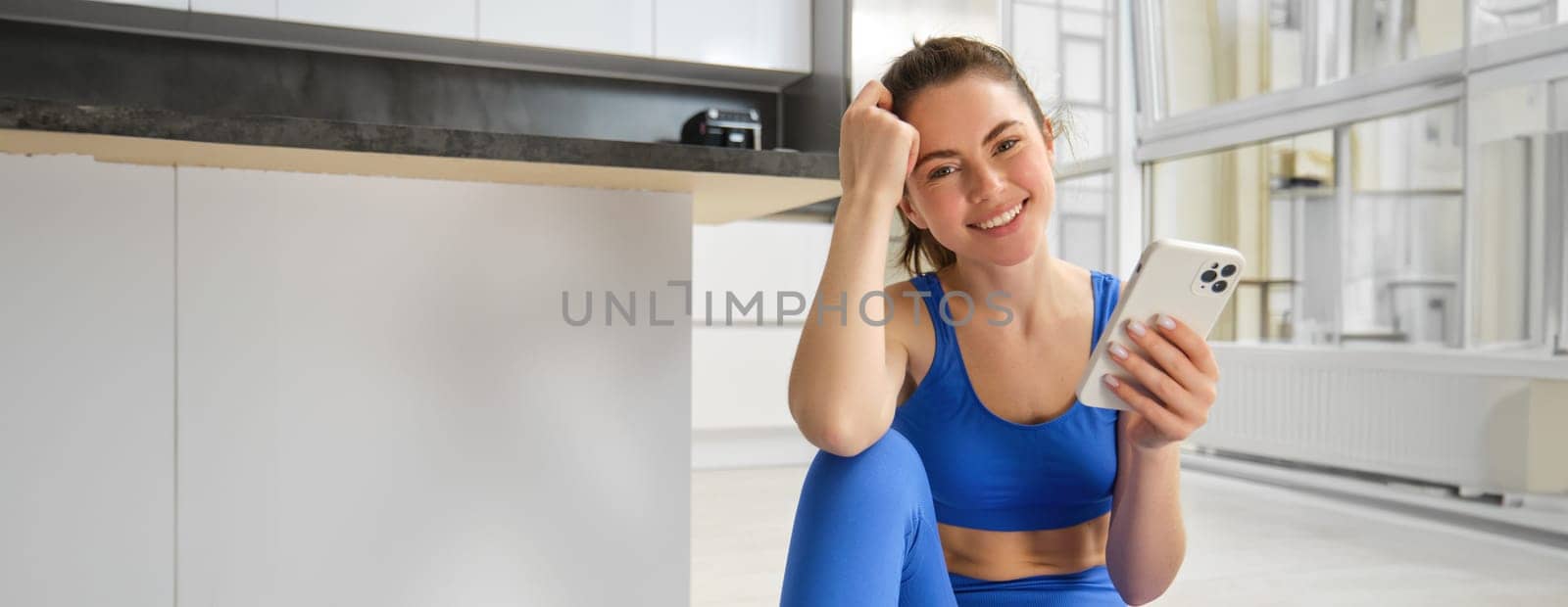 Young woman with smartphone does workout at home, using mobile phone app for sports training indoors, wears blue sportsbra and leggings.