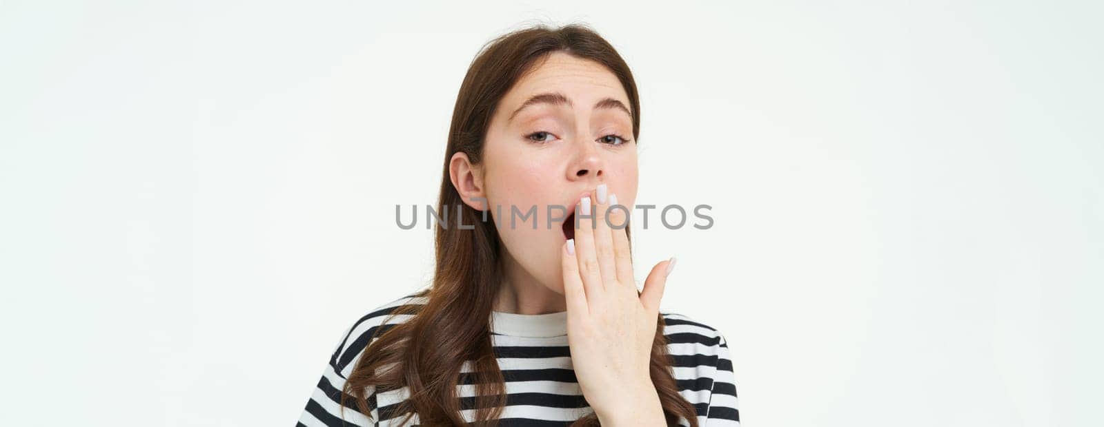 Portrait of girl yawning from boredom, covers her mouth with hand, looks sleepy, wakes up late, stands over white background.