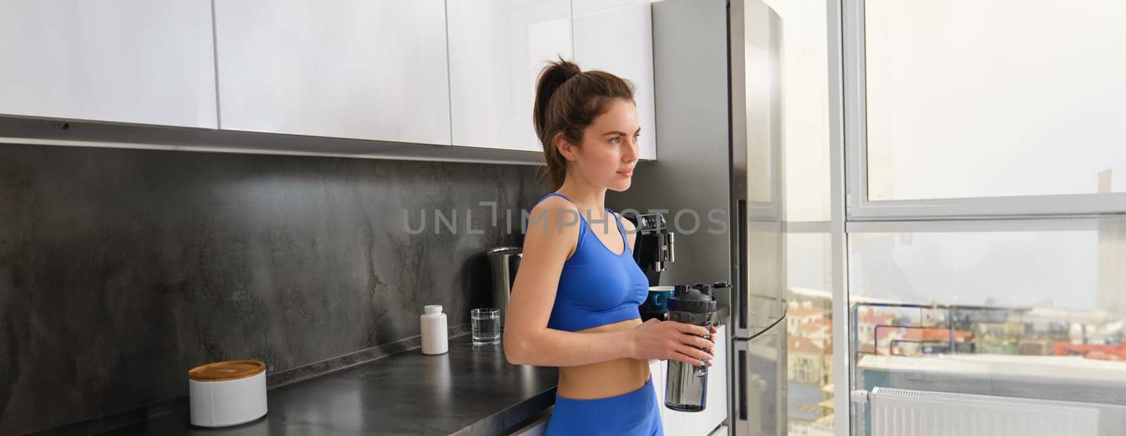 Image of fit and healthy, fitness woman in sportswear, drinking water, holding bottle shaker and standing in kitchen, staying hydrated after workout by Benzoix