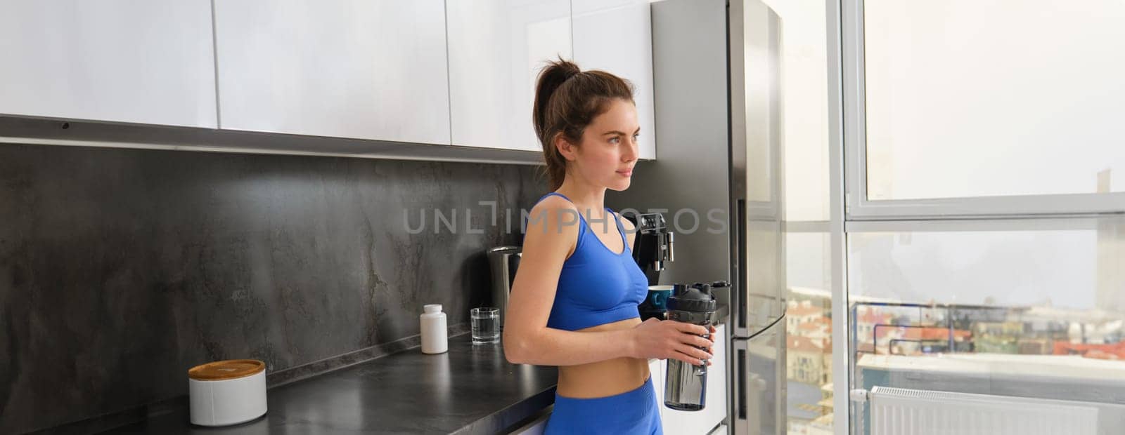 Image of fit and healthy, fitness woman in sportswear, drinking water, holding bottle shaker and standing in kitchen, staying hydrated after workout.