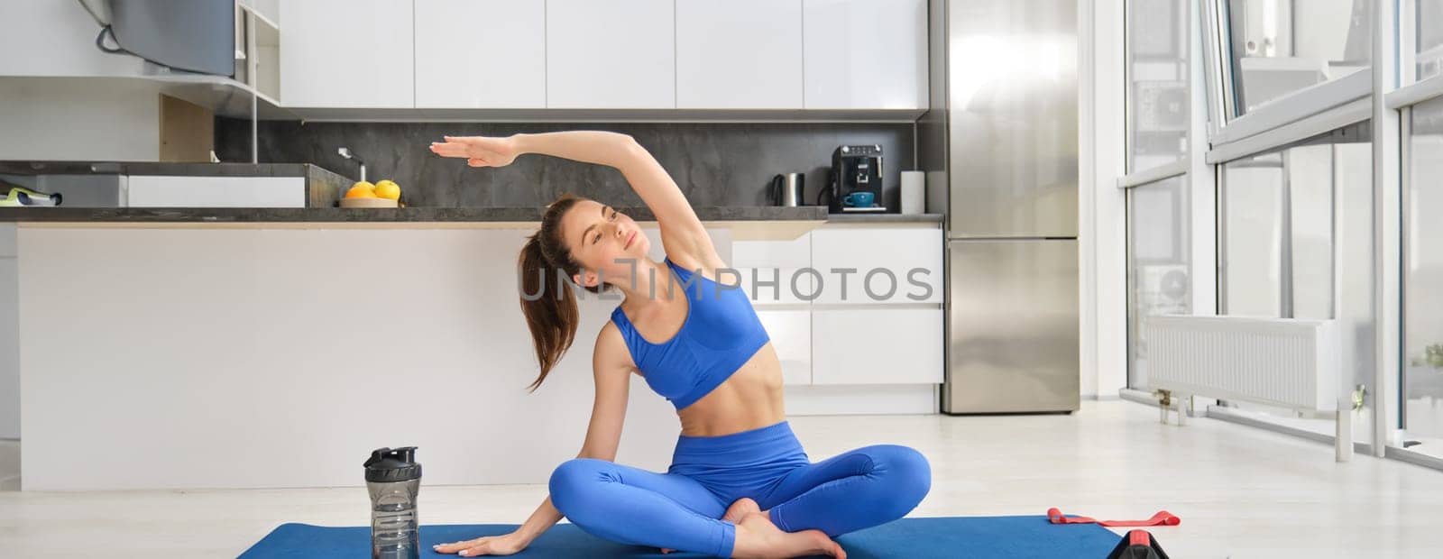 Portrait of sporty woman does fitness workout from home, sits on blue rubber yoga mat during training, stretching hands.
