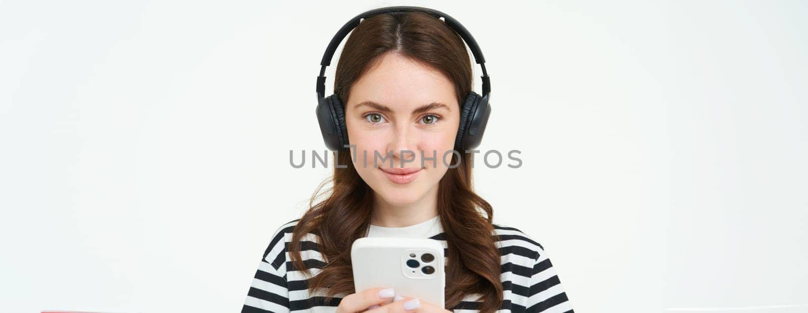 Image of brunette young woman, smiling, listening to music in headphones, watching videos on mobile phone app, holding smartphone, standing over white background.