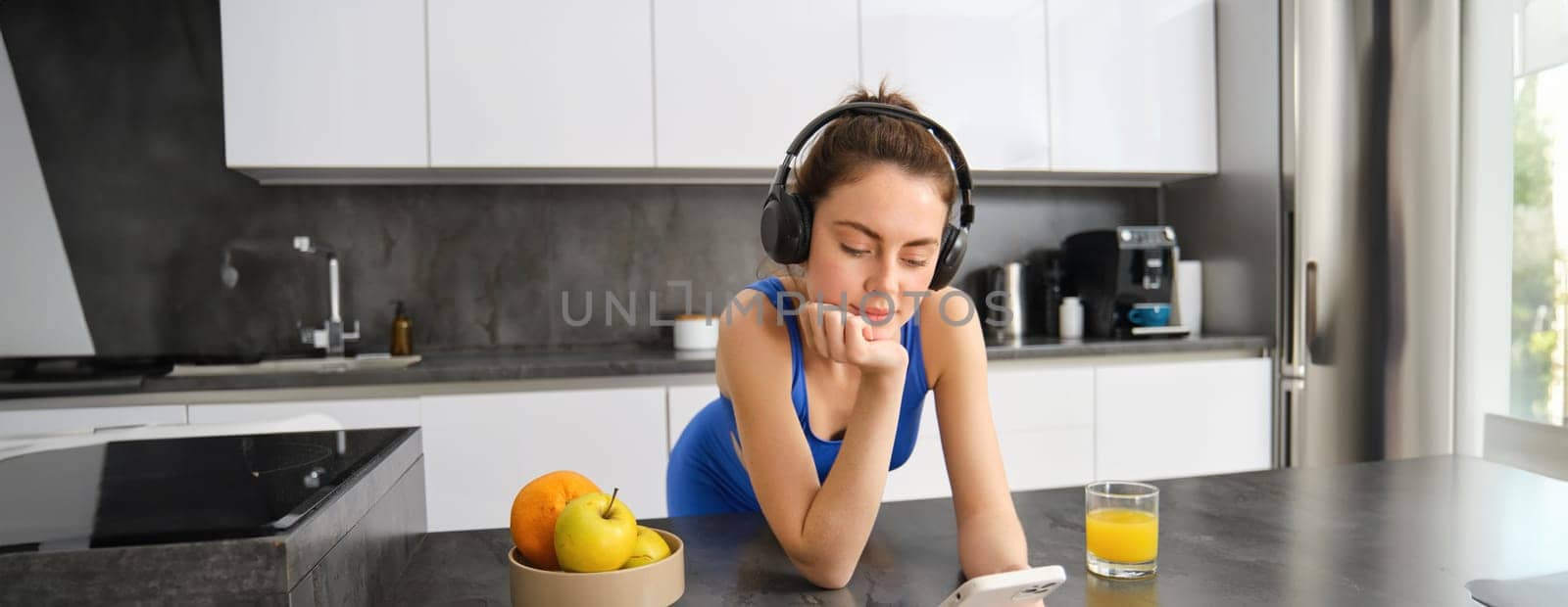 Portrait of young fitness woman with headphones, drinking orange juice in kitchen and using smartphone, listening music, getting ready for workout gym.